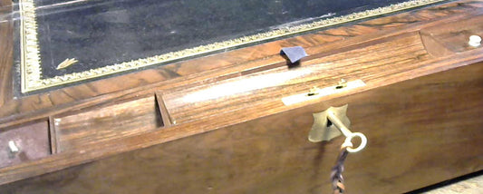 A wooden writing slope with a black leather top panel bordered by gold detailing, featuring brass hardware and a hanging key.